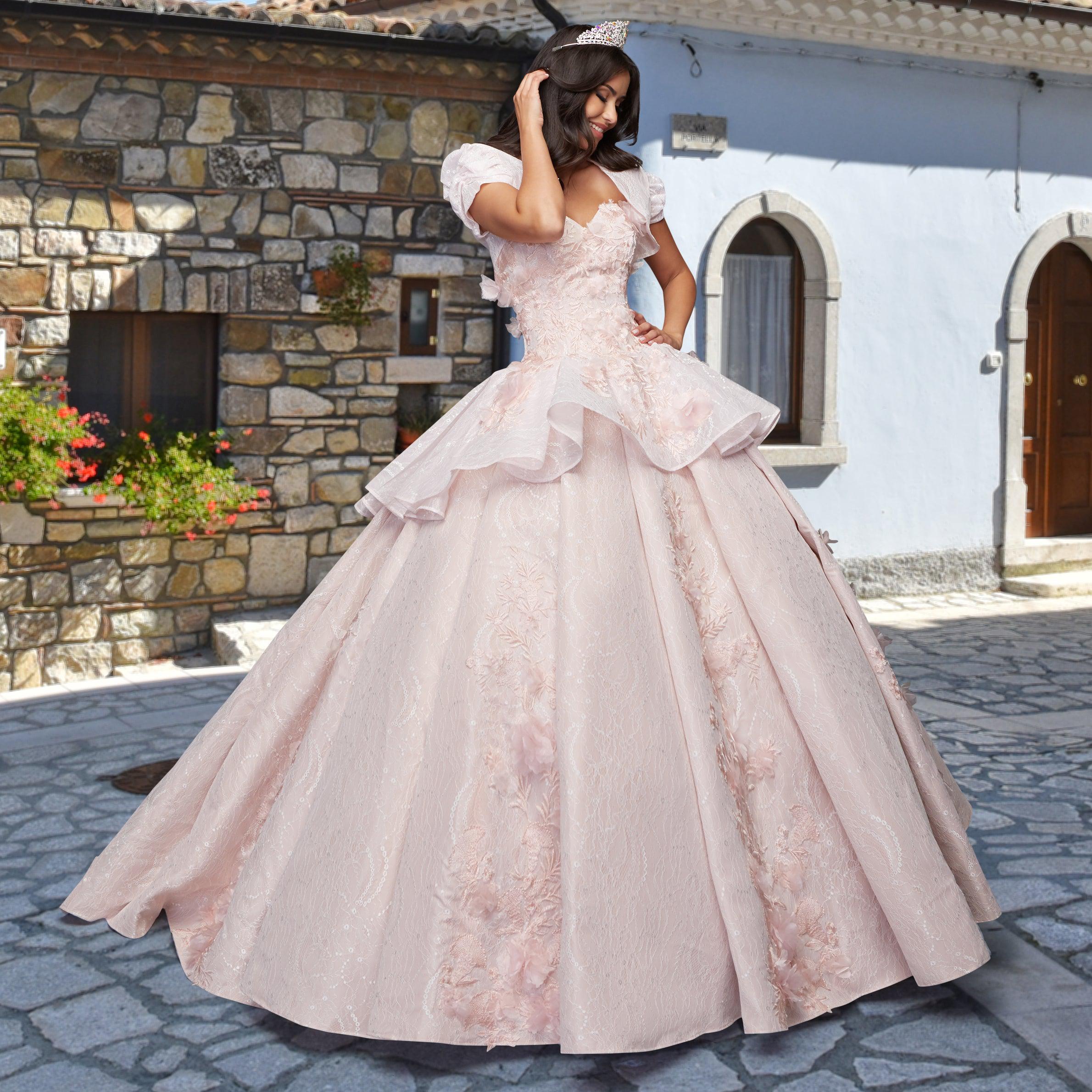 A woman wearing the Quinceañera Sample Dress QF115 and a tiara poses on a stone-paved street, set against rustic buildings with stone and plaster walls.